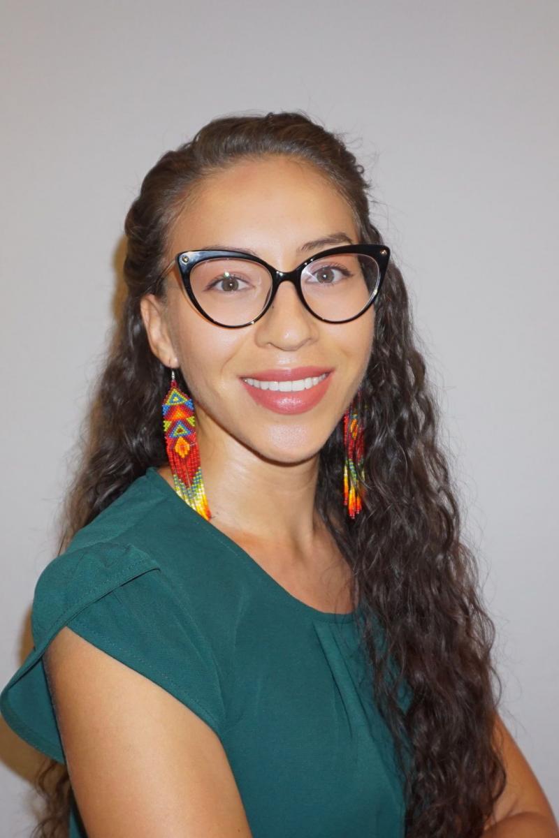 Dr. Dina Verdin in front of a white background wearing a dark teal, short-sleeved blouse, black thick-rimmed, wingtip glasses, and colorful long beaded earrings