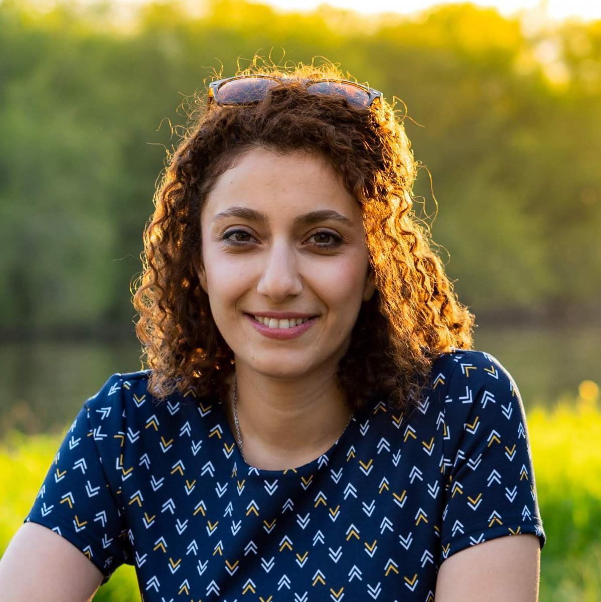 Dr. Mitra Asgari outside with out-of-focus greenery in the background weating a blue short-sleeved blouse with white and yellow chevrons and sunglasses on top of her head