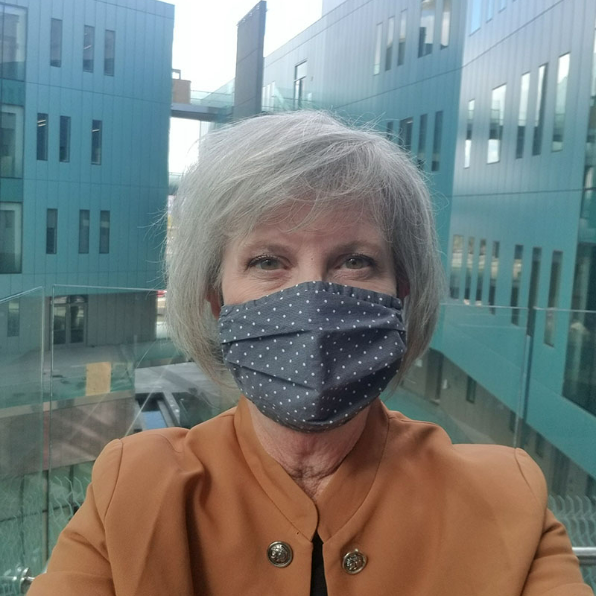 Amy Pate in front of a window overlooking blueish-grey modern buildings outside wearing a taupe top and a grey cloth facemask with small white polka dots