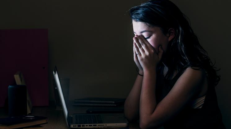Student sitting in the dark in front of their laptop looking dismayed with their face in their hands