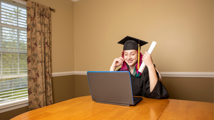 student with rainbow colored hair wearing graduation clothing at a computer.