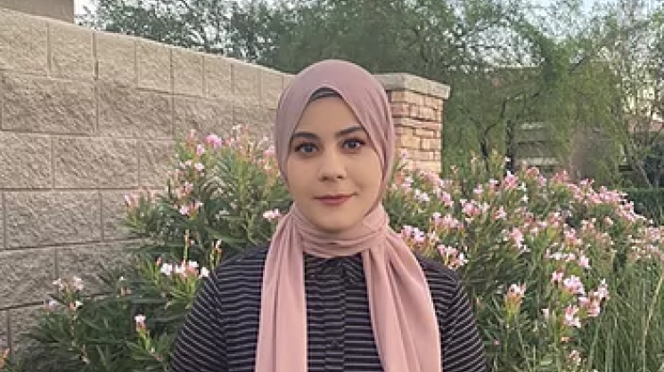 Photo of Tasneem Mohammed wearing a black and white horizontal striped shirt and a rose-colored hijab, standing outside in front of a green bush with pink flowers