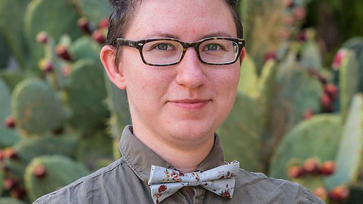 Headshot of Tay Misheva wearing a grey collared shirt and bowtie ad black-rimmed glasses, standing in front of a prickly pear cactus