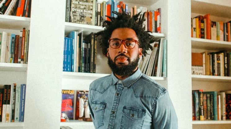 Dr. Keon McGuire wearing a button-up denim shirt and brown/red-rimmed glasses standing in front of white built-in bookshelves filled with books