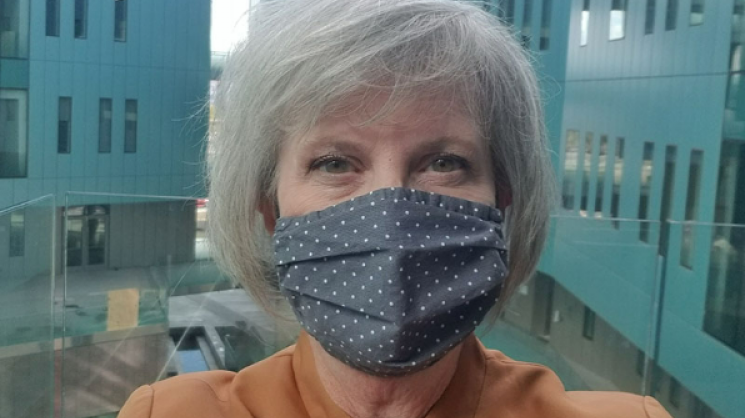 Amy Pate in front of a window overlooking blueish-grey modern buildings outside wearing a taupe top and a grey cloth facemask with small white polka dots