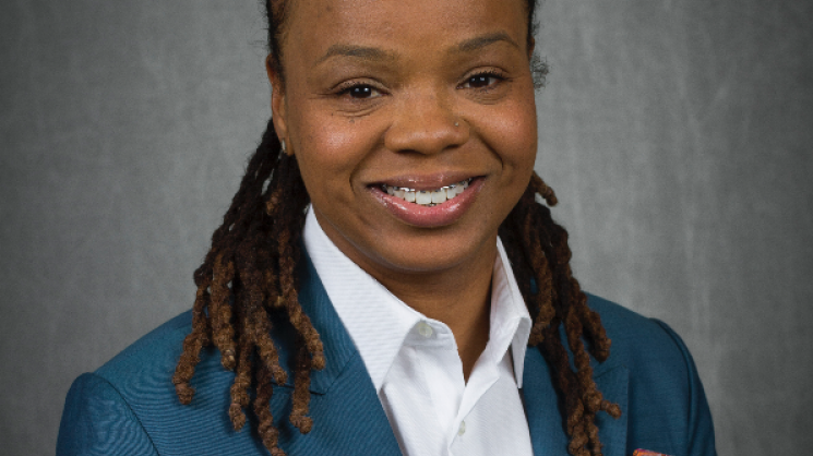 Headshot of Dr. Brooke Coley in front of a grey background wearing a white button-up shirt and blue blazer with a colorful pocket square
